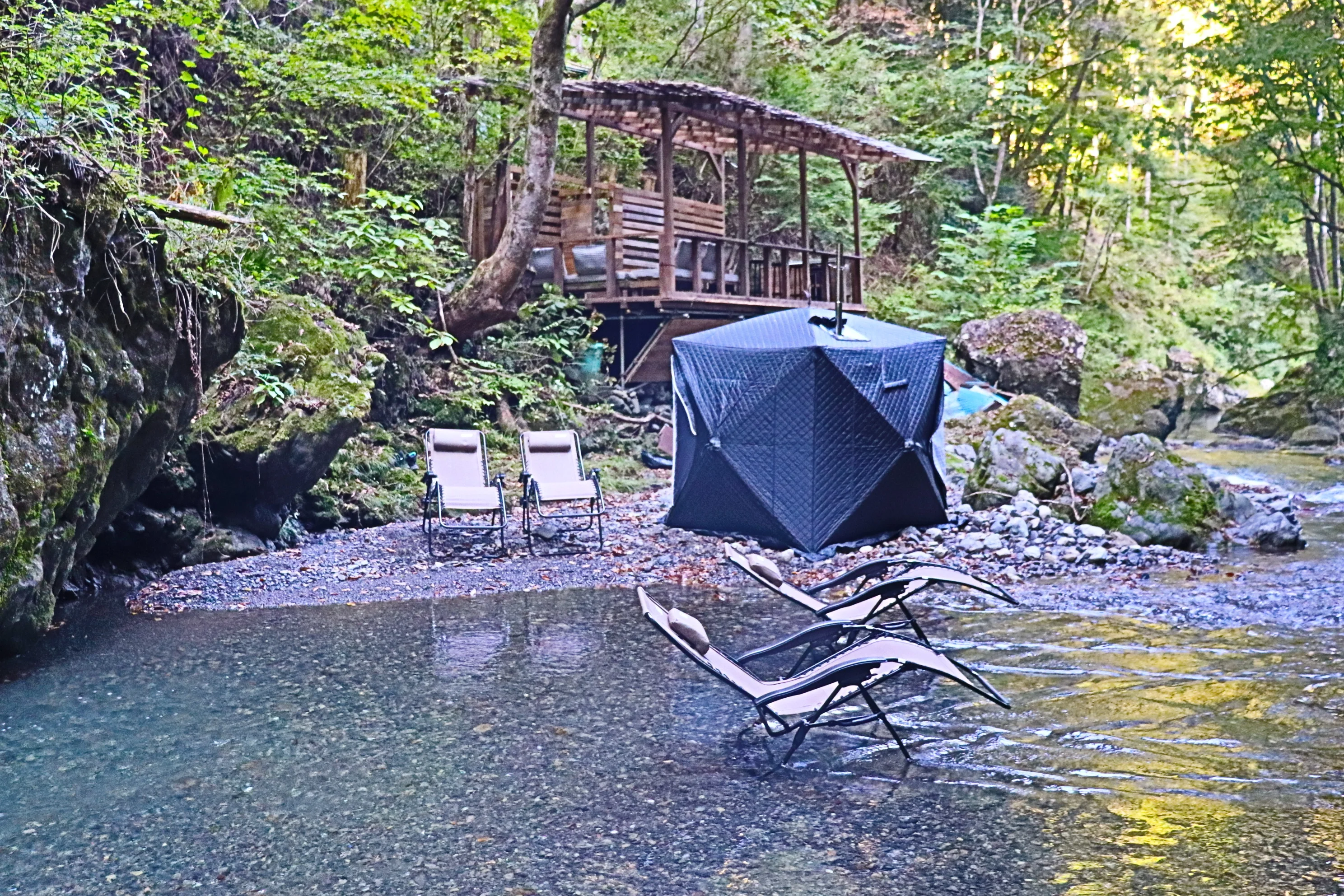 川辺の屋根付きデッキ小屋と黒いテントサウナ、川の中のリクライニングチェア